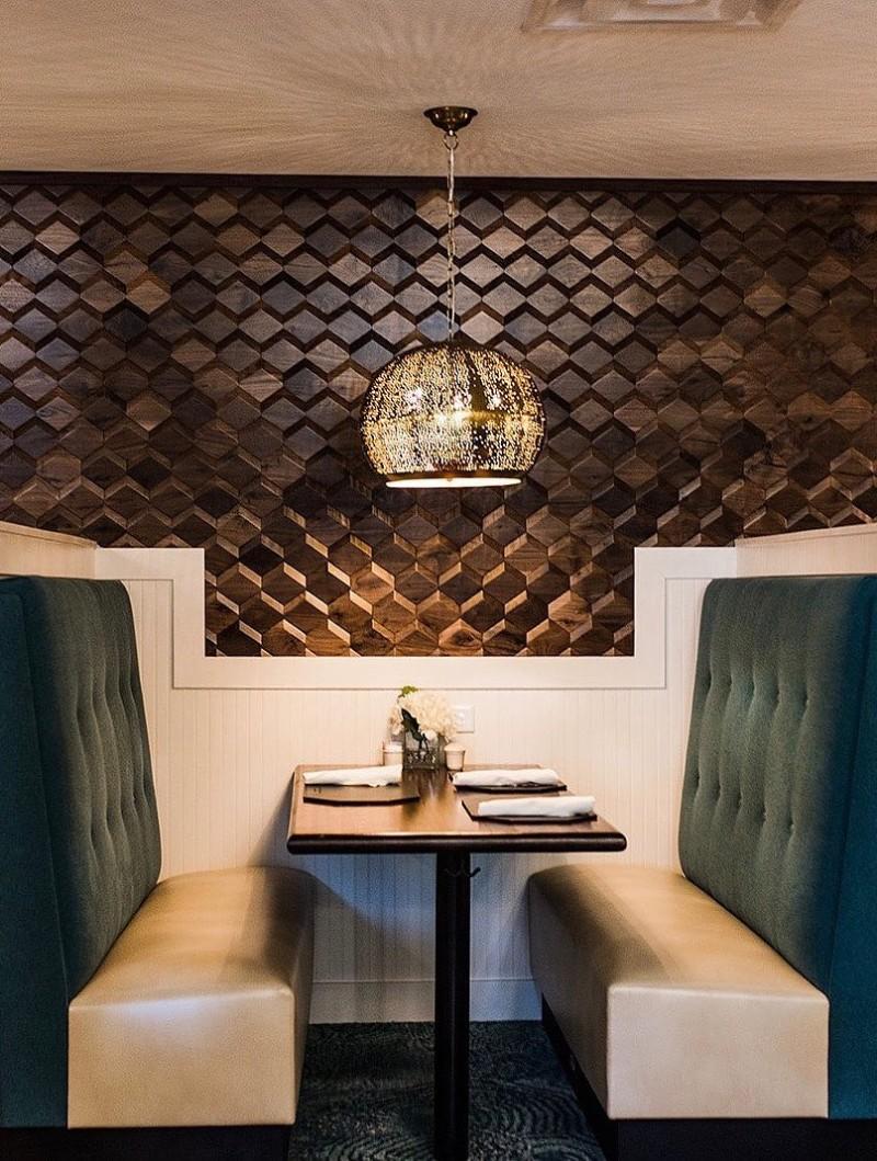 Restaurant booth with green backed benches, a brown designed wooden wall, and a natural brass pierced chandelier