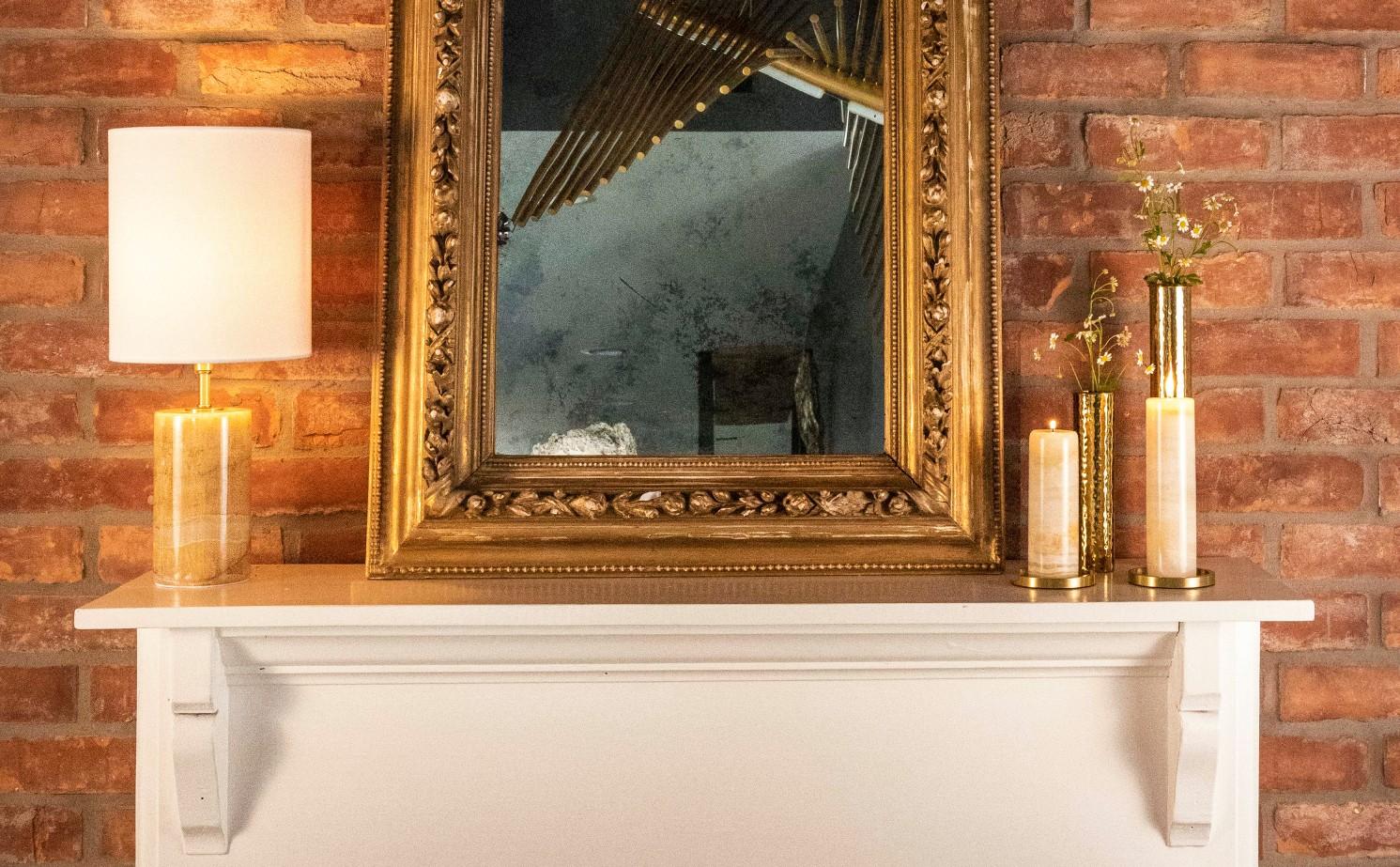 White mantel in front of a red brick wall with an ornate mirror, jade lamp, jade candle holders, and hammered natural brass caandle holders