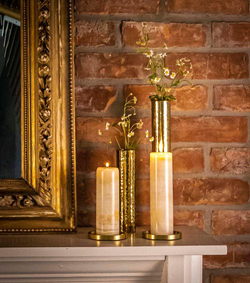 White mantle with red brick wall, ornate gold frame, jade candle holder, and hammered natural brass candle holder