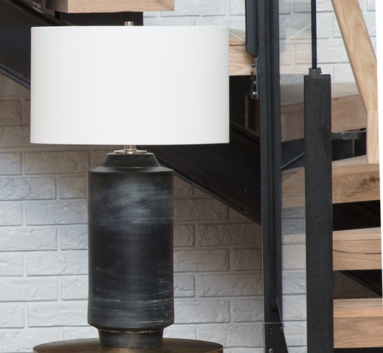 Black cylindrical ceramic table lamp on a small side table in front of a spiral staircase