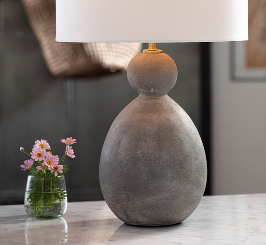 Gray ceramic table lamp with a flared base and bulbous top next to a cup with water and flowers