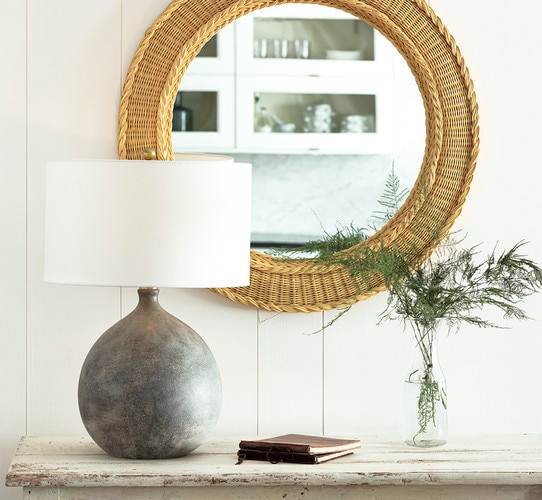 Circular gray ceramic lamp on a rustic white console table in front of a wicker lamp