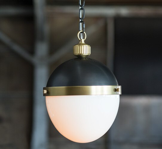 Brass and black pendant light with a milk glass shade in front of a metal and wood wall