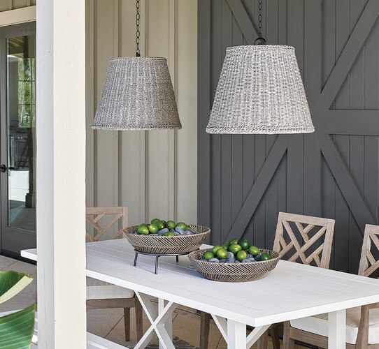White outdoor dining table with two baskets of limes and two gray woven pendant lights above it