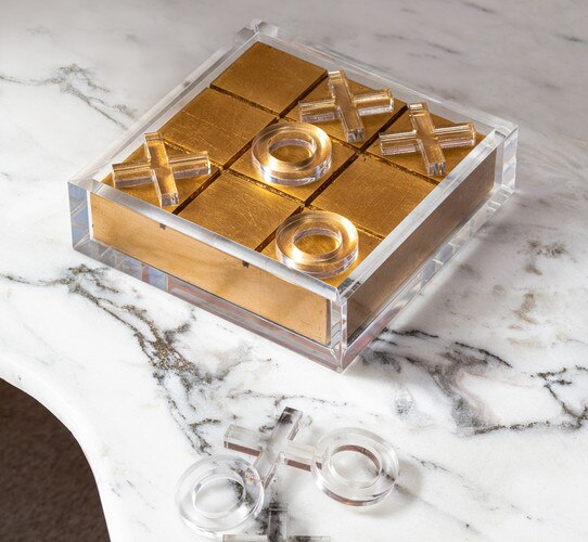 Gold and acrylic tic tac toe board with clear acrylic pieces on a marble countertop