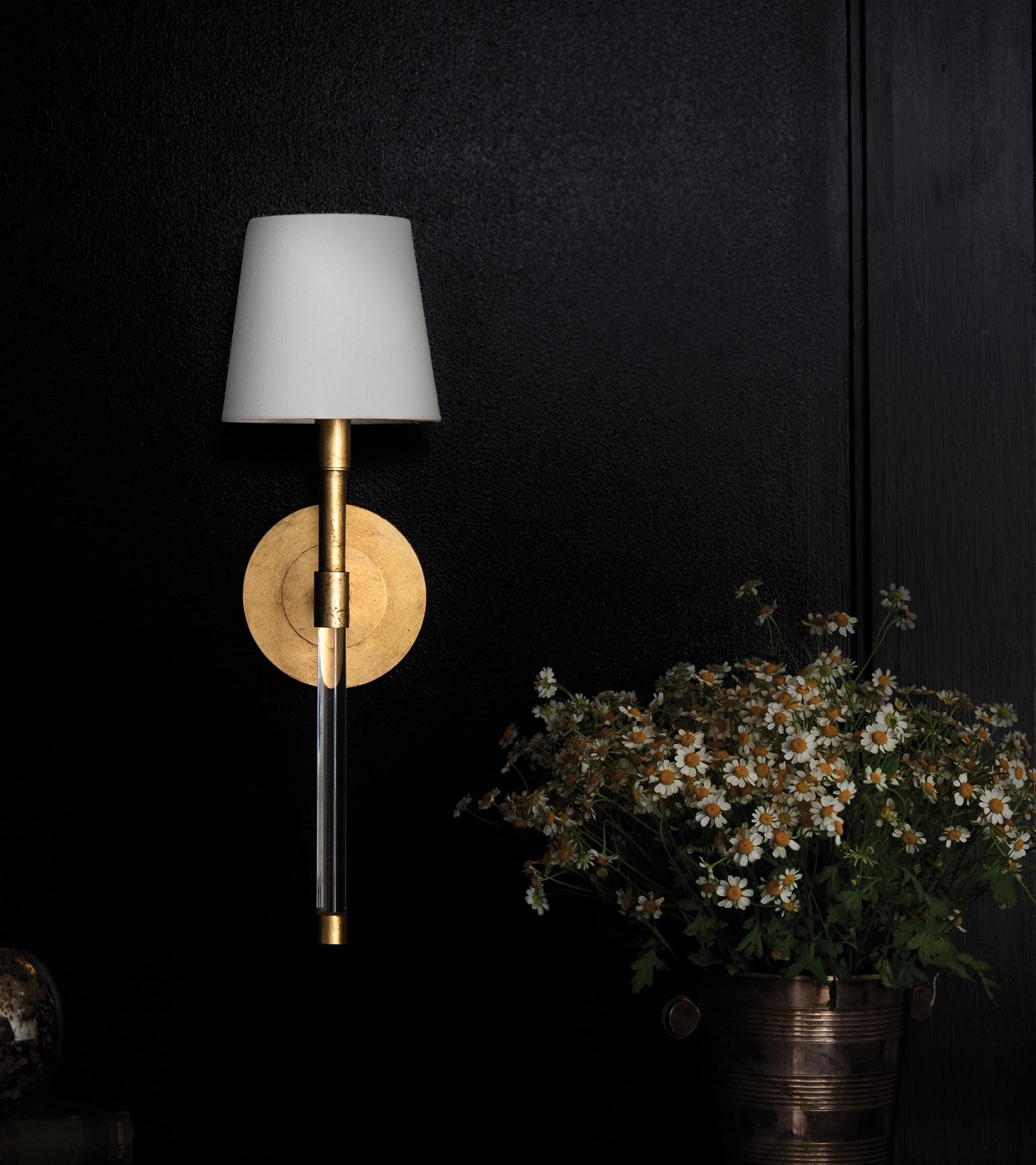 A natural brass sconce with a white shade on a dark wall colored wall next to a pot of flowers